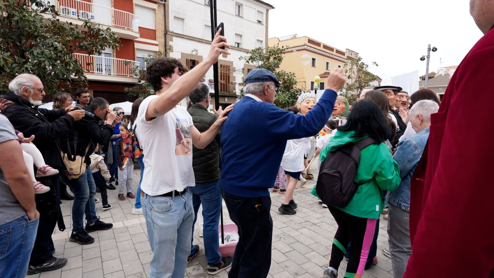 En una plaça, un grup de persones es reuneixen amb gegants, mentre altres fan fotos i graven vídeos amb els seus telèfons mòbils. Al fons, es poden veure edificis de diversos pisos.