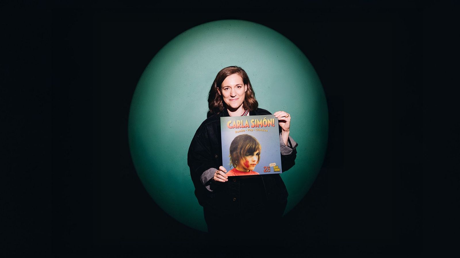 Carla Simón, amb cabells castanys ondulats i una jaqueta fosca, sosté una portada amb el seu nom i el títol "VERANO 1993", acompanyada per la imatge d'una nena.