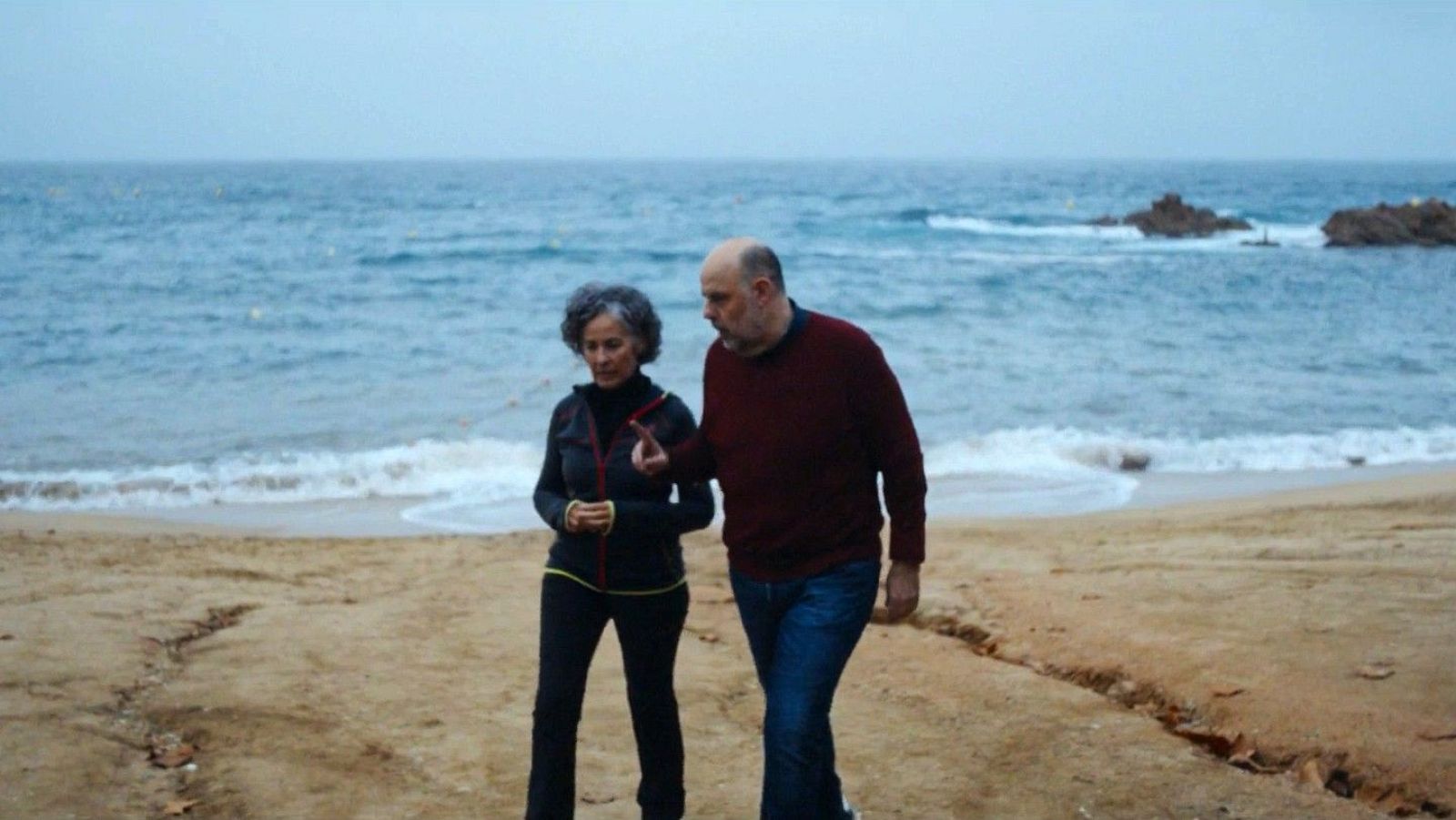 Jordi Basté i Nina caminen per la platja; ella amb jaqueta fosca i detalls grocs, ell amb jersei granat. El mar, amb onades, completa l'escena.