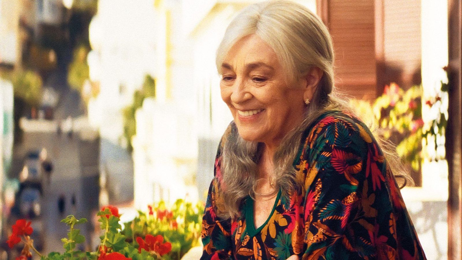 Carmen Maura, con una sonrisa, posa en un balcón con flores rojas, luciendo un vestido con estampado floral, con una calle y edificios al fondo.