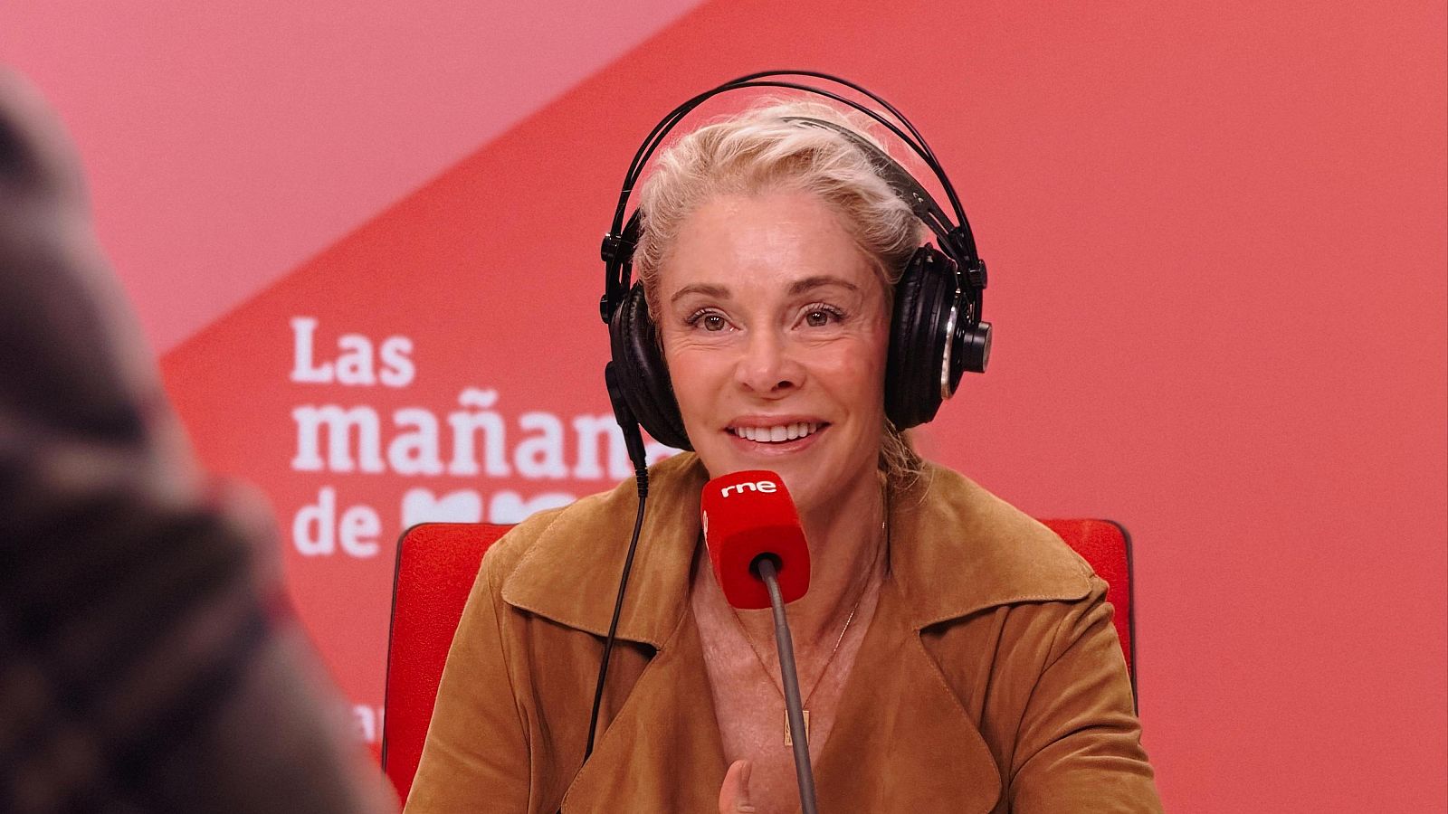 Belén Rueda, con el pelo rubio recogido y auriculares, sonríe en un estudio de radio frente a un micrófono rojo. Se encuentra vestida con una chaqueta marrón, con el logo de RNE y el nombre de un programa de radio al fondo.