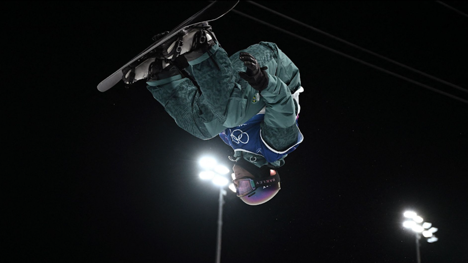 Una esportista de snowboard realitza un salt acrobàtic, invertida amb la taula. Vesteix amb roba verda i porta casc i ulleres protectores. El fons, fosc, està il·luminat per focus.