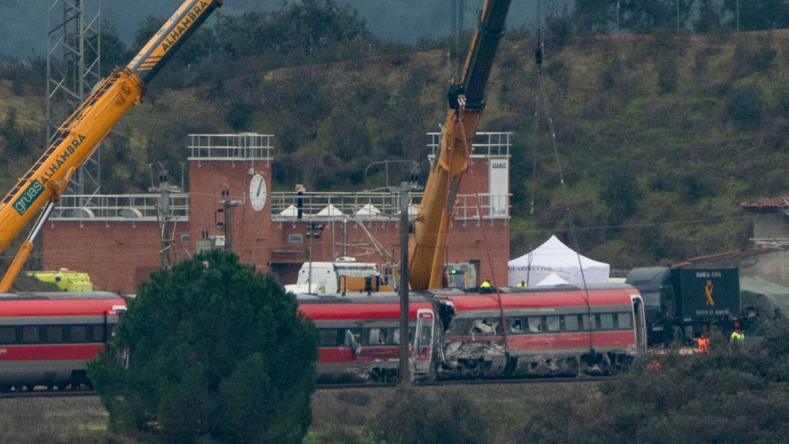 Un tren rojo dañado y descarrilado es el foco de atención, con dos grúas amarillas operando en el lugar. Se observa un edificio de ladrillo con un reloj y la presencia de personal de emergencia.