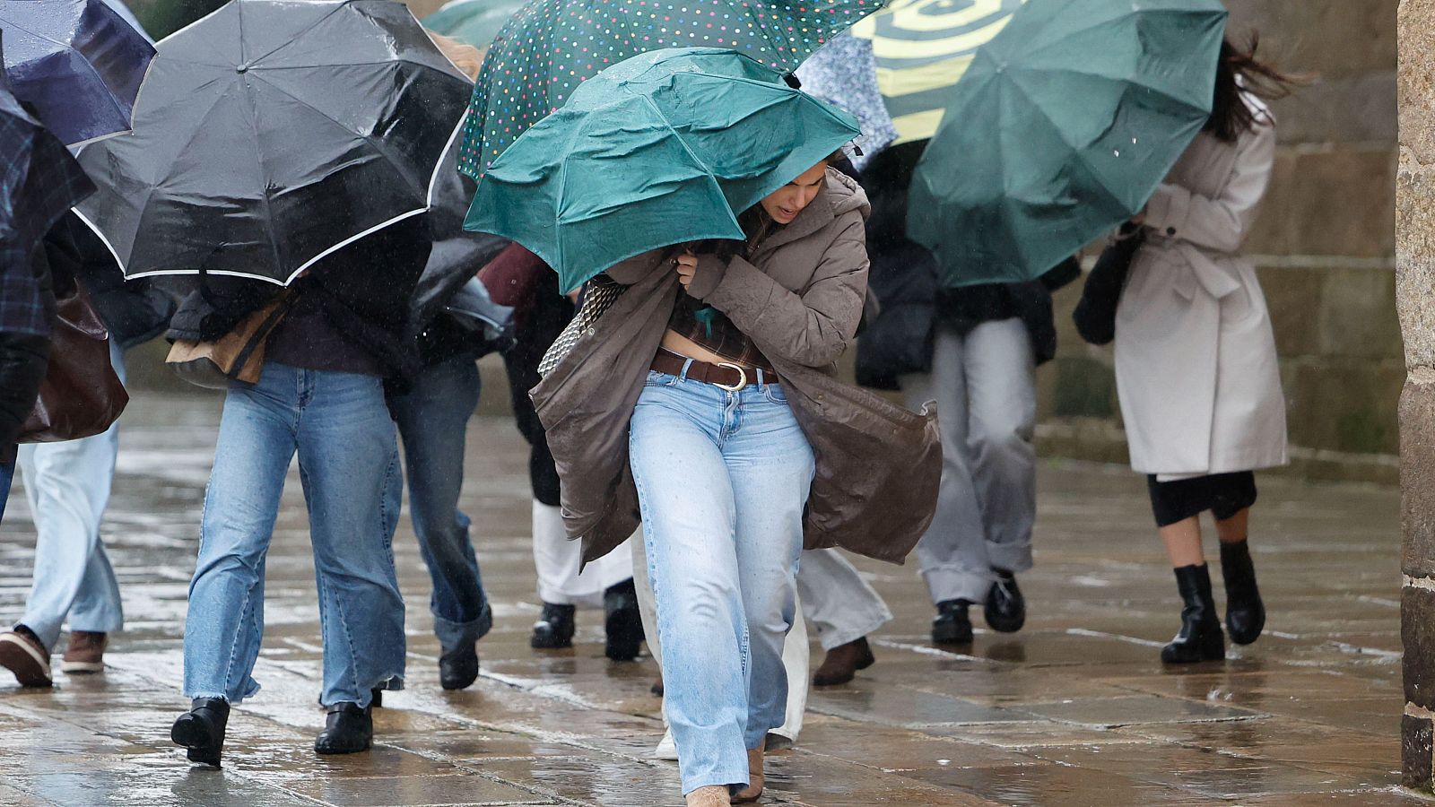 En una calle de Galicia, un grupo de personas se resguarda de la lluvia y el viento con paraguas de distintos colores. Se aprecia a una mujer con abrigo marrón y vaqueros, mientras el suelo mojado refleja la luz.