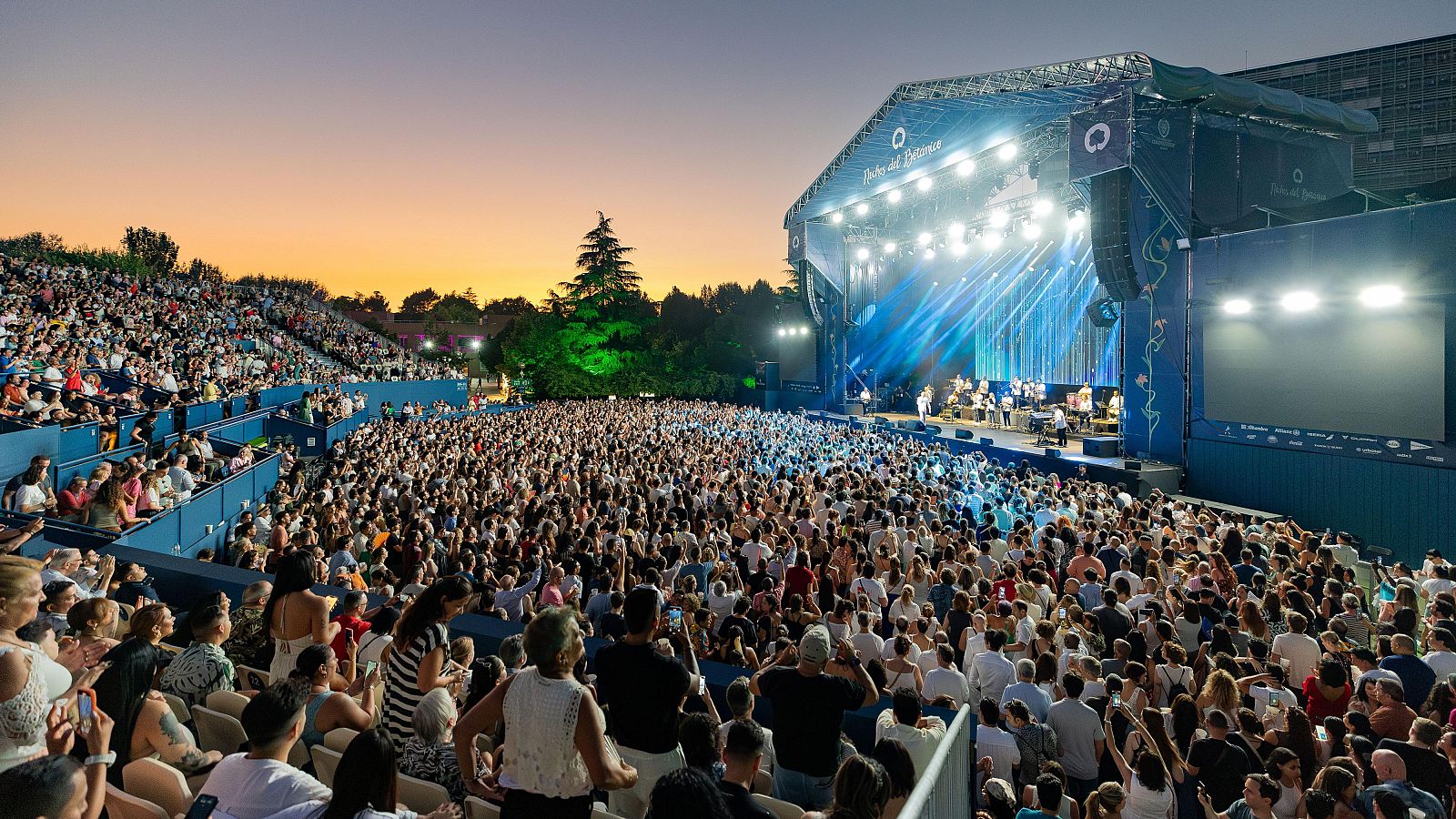 Un escenario iluminado con una banda musical en acción, frente a una multitud entusiasta. El evento al aire libre se desarrolla al atardecer, con una pantalla gigante y un ambiente festivo.