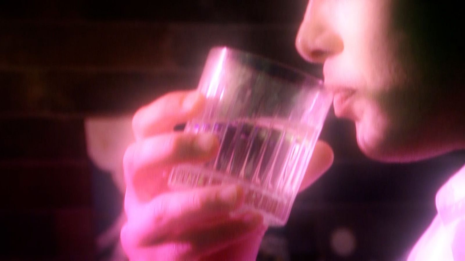 Un hombre bebe de un vaso transparente en un entorno con iluminación tenue y tonalidades rosadas, posiblemente en un ambiente íntimo.