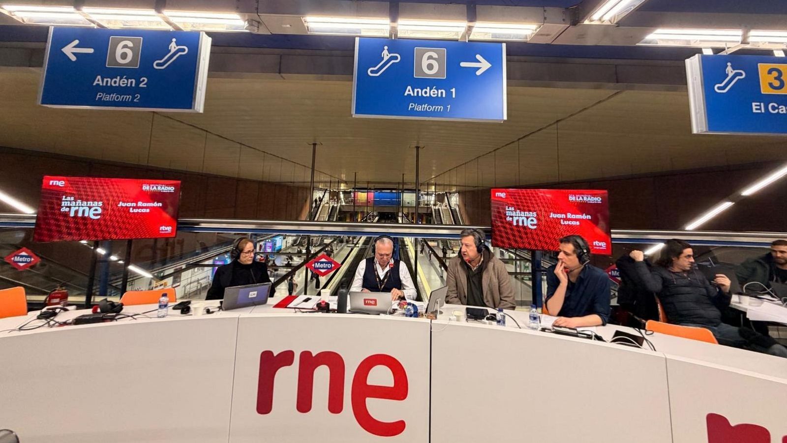 Una retransmisión de radio se lleva a cabo en una estación de metro, con un grupo de personas sentadas tras una mesa con el logo de la emisora, equipadas con micrófonos y auriculares. Se observan carteles indicadores y escaleras mecánicas en el fondo.