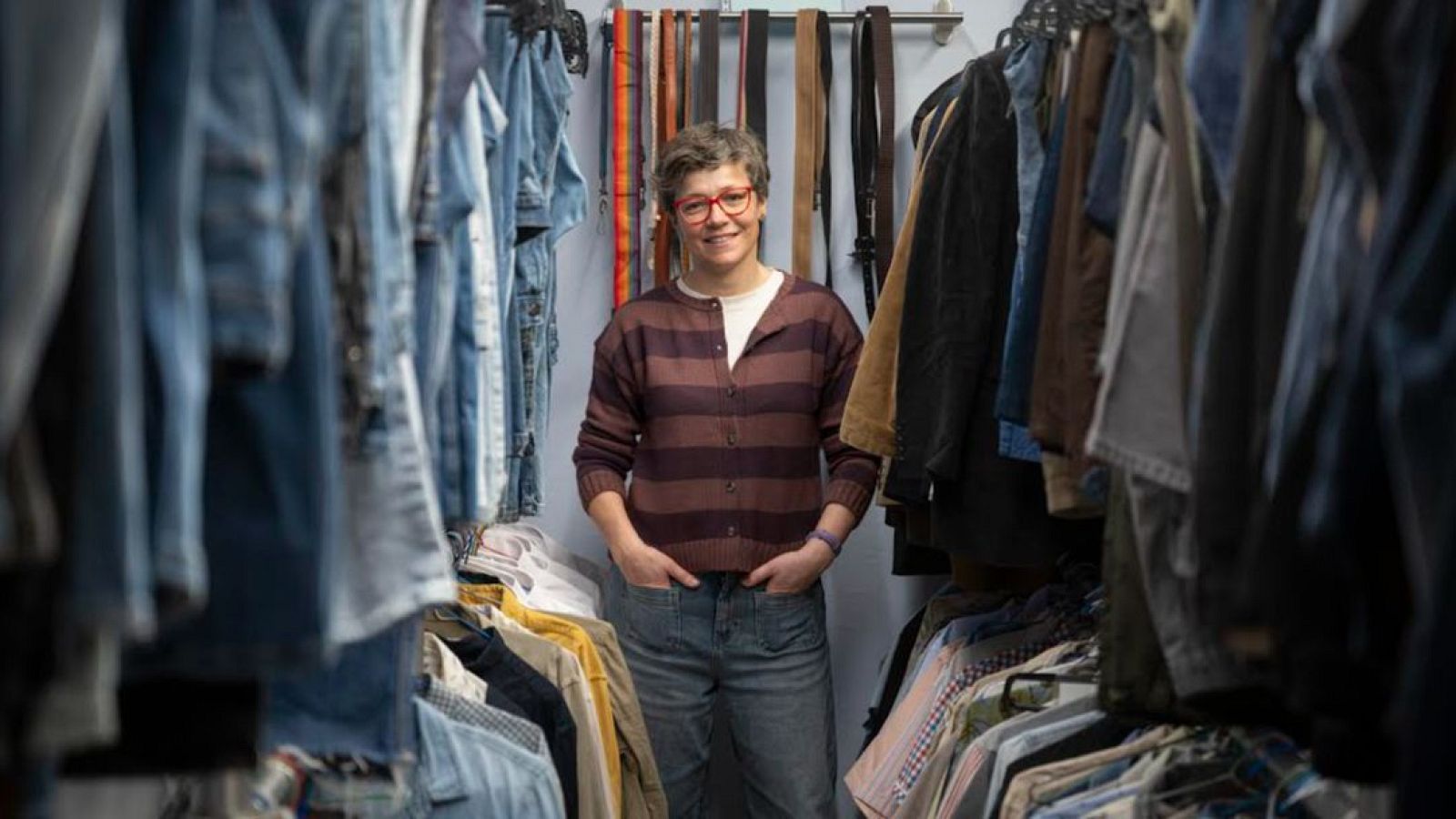 Bea Fernández, directora de la Fundació Arrels, apareix en una imatge envoltada de roba penjada. Vesteix una jaqueta de punt amb ratlles, texans i una pulsera, amb les mans a les butxaques.