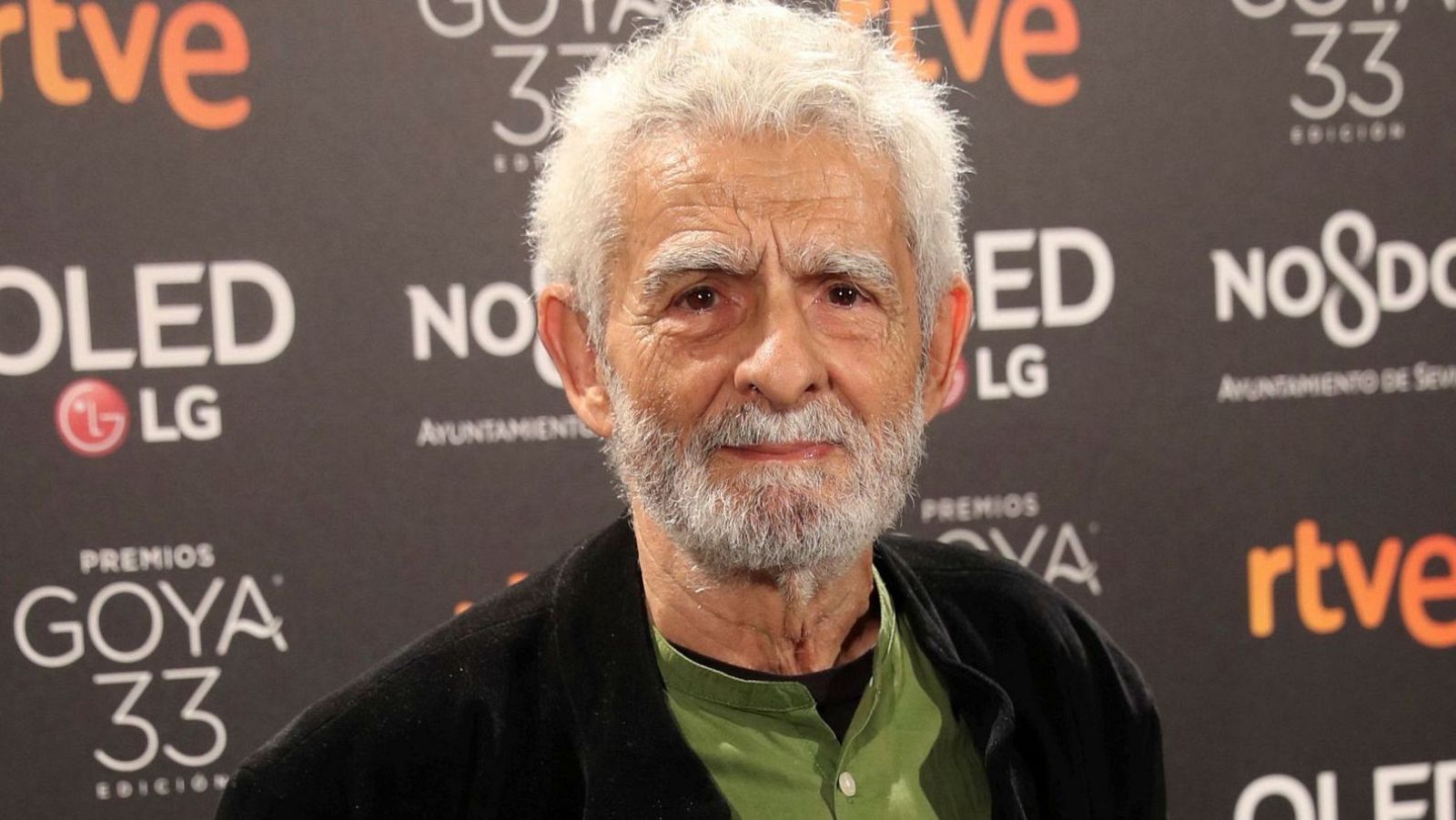 Un primer plano muestra a un hombre mayor con barba y cabello blancos, con una camisa verde y chaqueta negra, posando frente a un panel con logotipos y textos.
