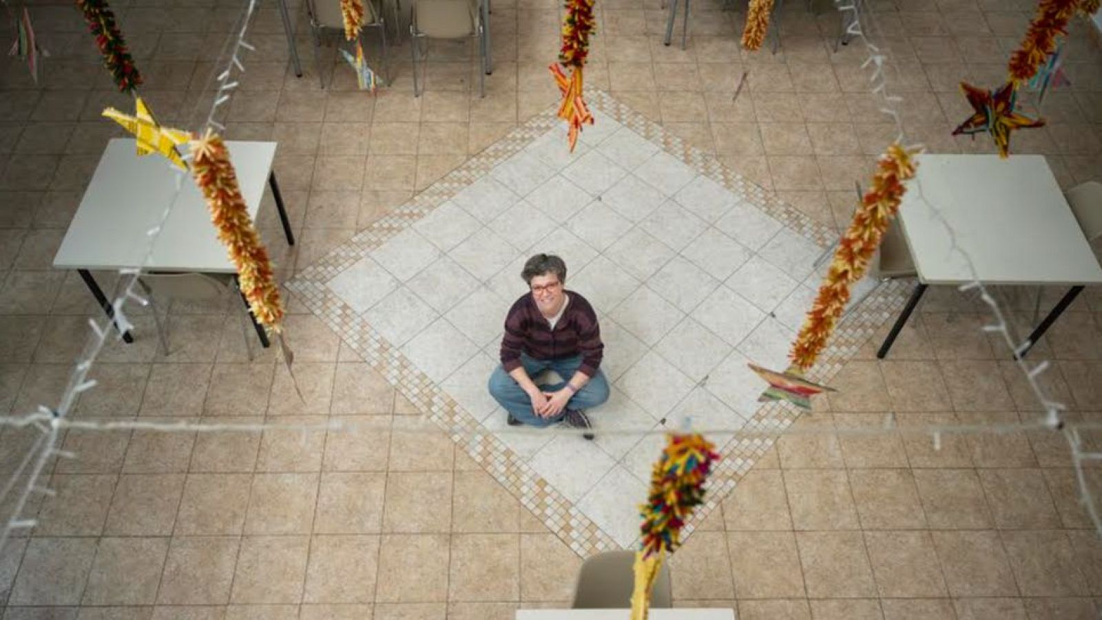 Una dona amb ulleres i camisa de quadres, presumiblement Bea Fernández, està asseguda al terra amb les cames creuades, envoltada de taules blanques i cadires transparents, amb guirnaldes de colors penjant del sostre.