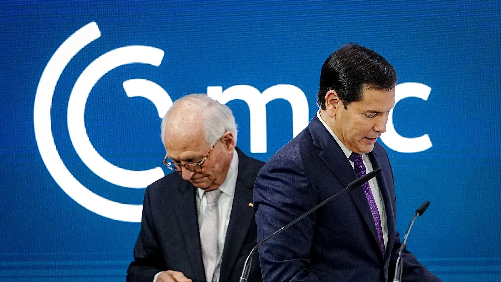 Dos hombres, uno de ellos con traje azul oscuro y corbata morada, se encuentran frente a micrófonos en un evento formal, con un fondo azul brillante y un logo blanco.