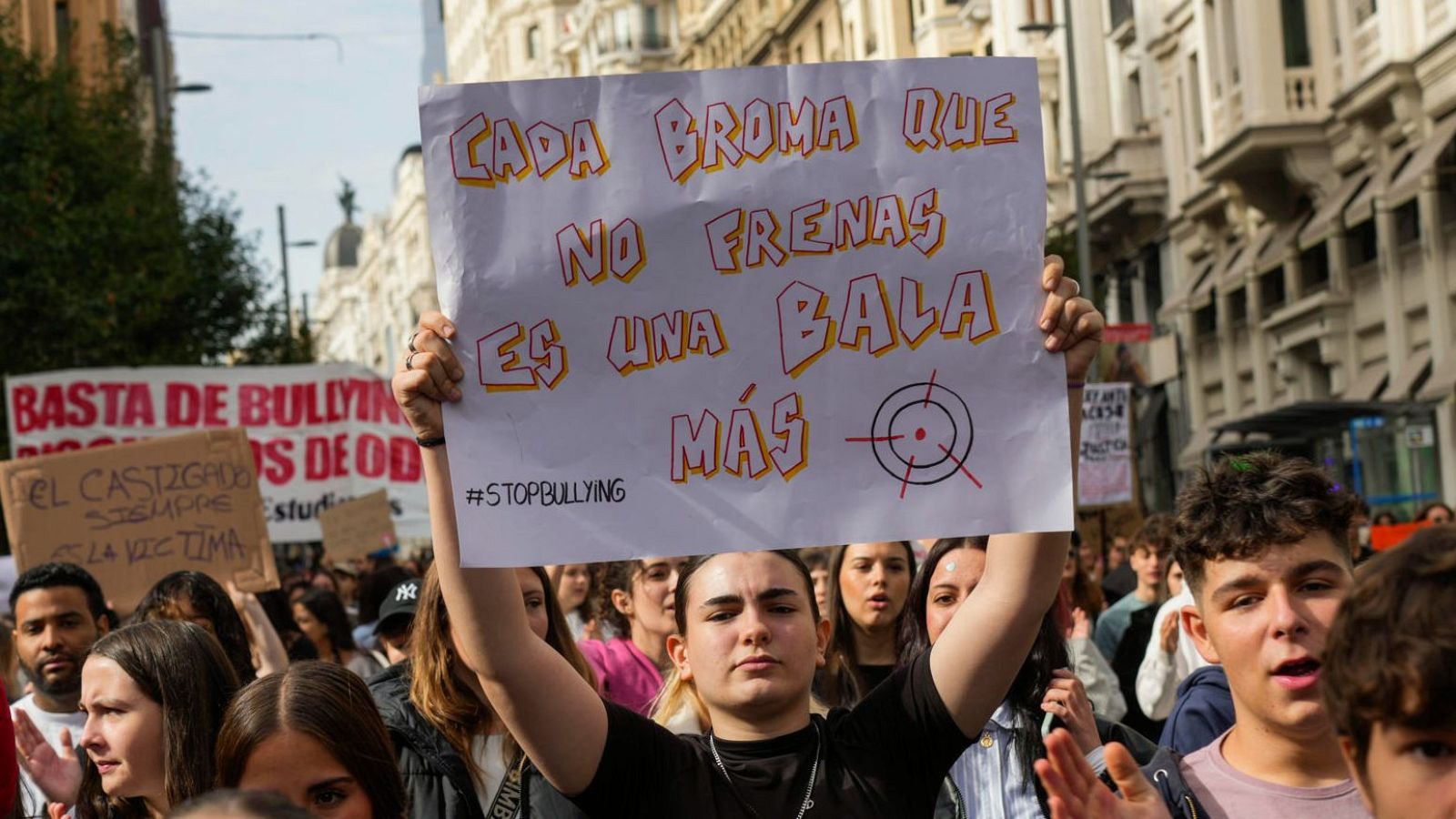 Una persona sostiene una pancarta con el mensaje "CADA BROMA QUE NO FRENAS ES UNA BALA MÁS" en una manifestación. Se observan más pancartas con mensajes contra el acoso escolar y un grupo de personas en una calle.