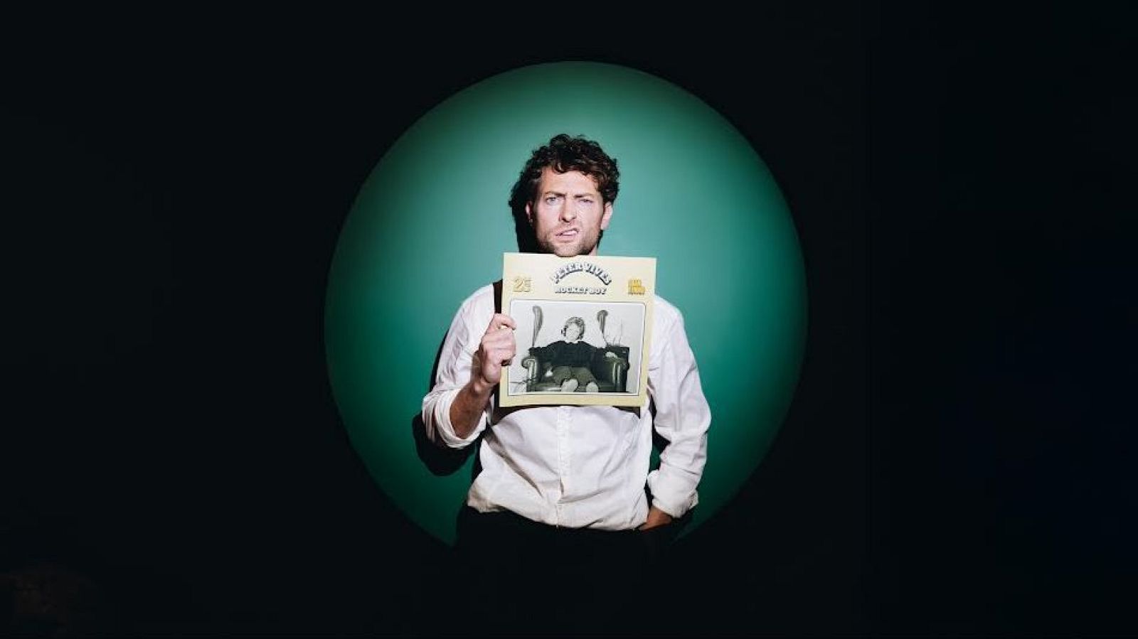 Peter Vives, amb camisa blanca i pantalons foscos, sosté un disc de jazz amb una portada en blanc i negre, il·luminat per un cercle verd sobre un fons negre.
