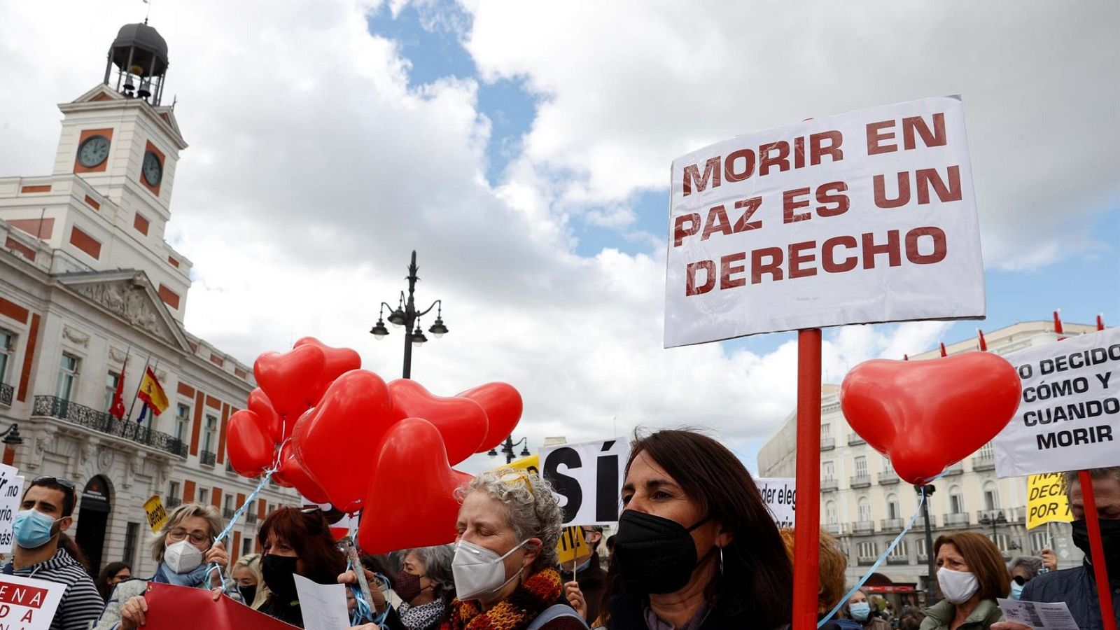 Una manifestación en la Puerta del Sol muestra a personas con mascarillas portando pancartas y globos rojos. Las pancartas expresan mensajes relacionados con el derecho a decidir sobre la propia vida.