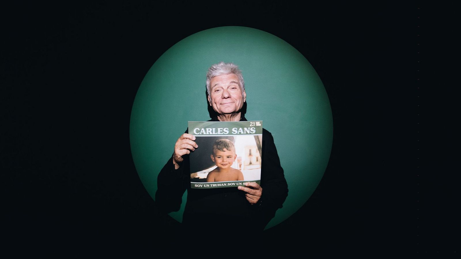 Carles Sans, con camisa oscura, muestra un disco cuya portada presenta una foto suya de niño y su nombre. El fondo es negro con un círculo verde claro.