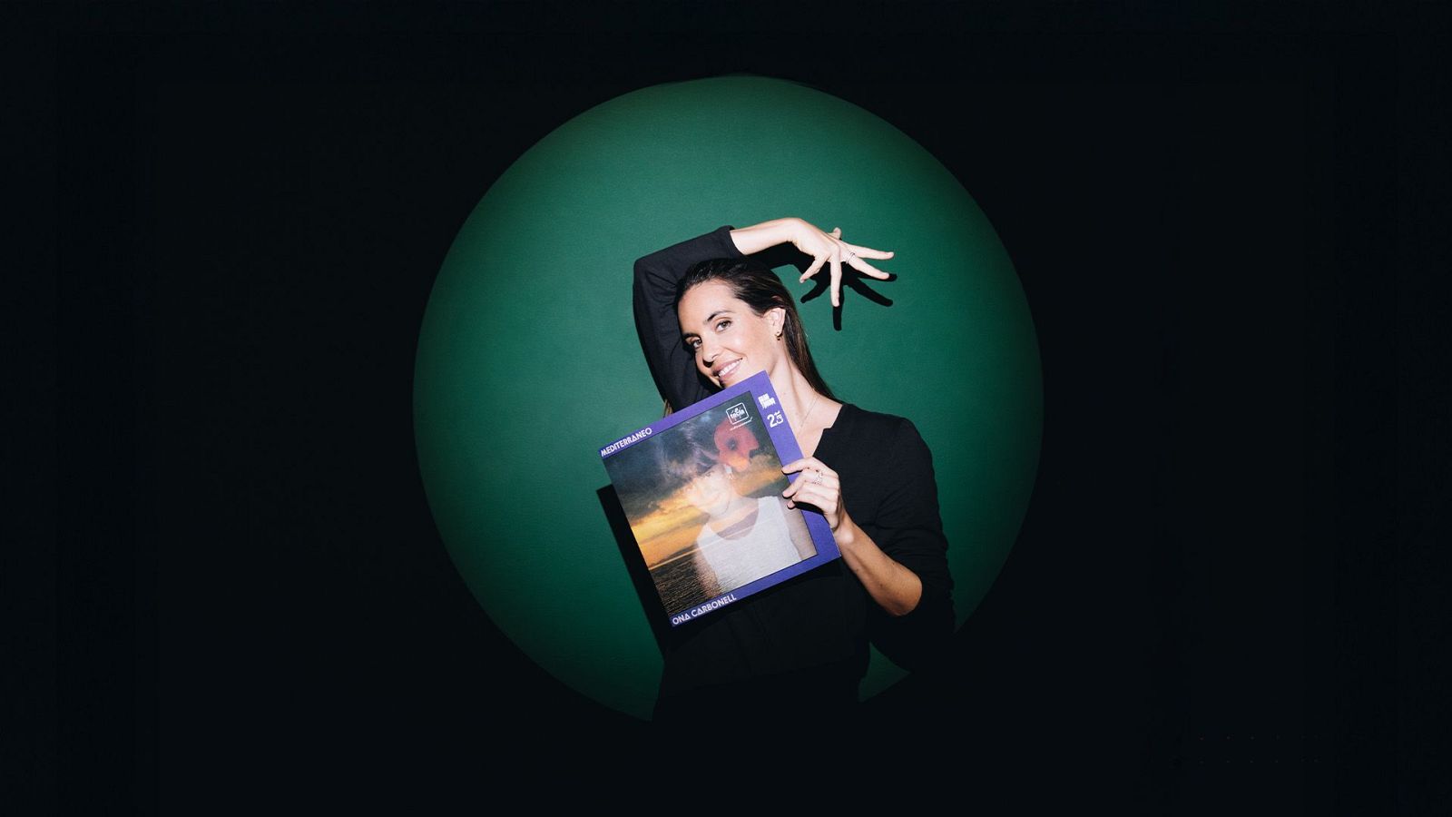 Ona Carbonell, amb cabells castanys i una blusa negra, sosté un disc amb la portada en tons blaus i taronges, on es llegeix el seu nom i "Mediterráneo".