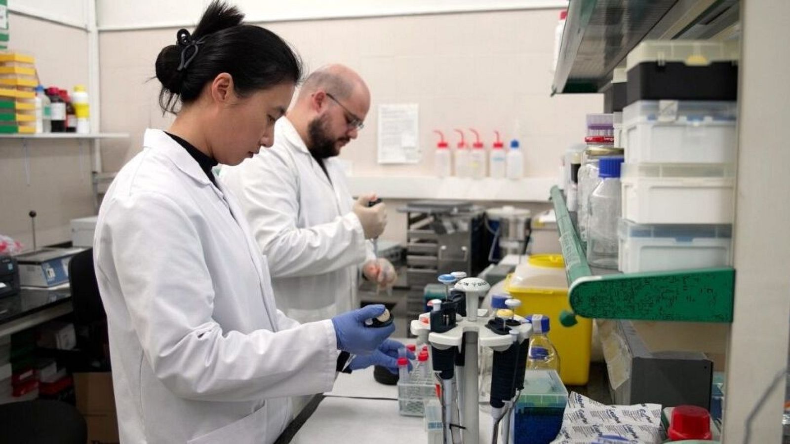 Dos científicos, una mujer con el pelo recogido y un hombre con barba y gafas, realizan experimentos en un laboratorio equipado con mesas y diversos instrumentos, ambos con batas blancas.