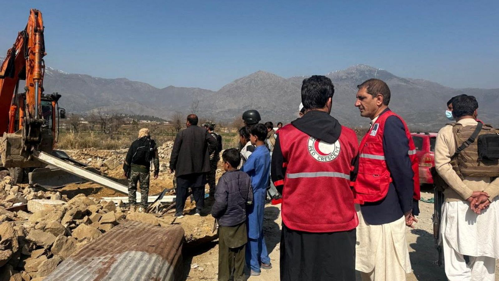 Una excavadora naranja trabaja entre escombros en una zona montañosa, con trabajadores de emergencia con chalecos rojos y civiles presentes. El cielo despejado y el paisaje montañoso completan la escena de respuesta a una emergencia.
