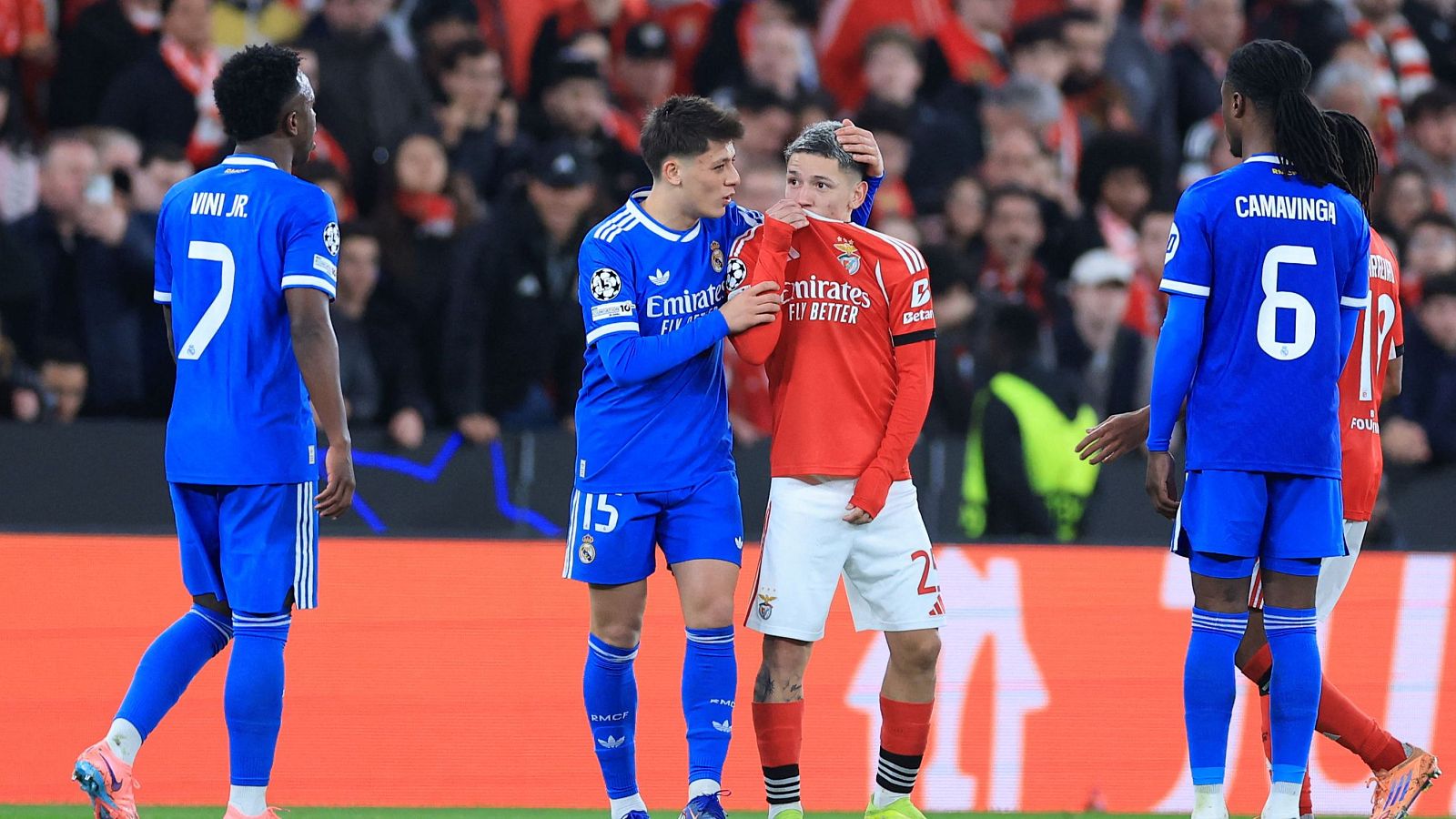 Vinicius Jr., con la camiseta número 7, y Prestianni, con la camiseta roja, interactúan en el campo de juego. Prestianni cubre su rostro, mientras Camavinga observa la escena de espaldas.