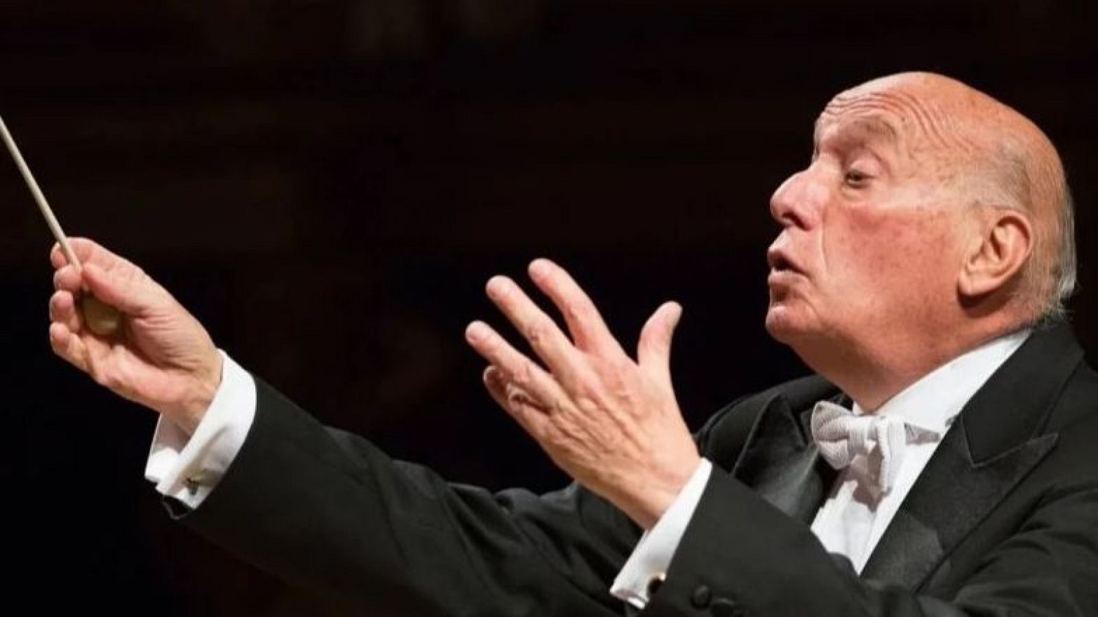 Pinchas Steinberg, con un esmoquin y pajarita, dirige con una batuta, concentrado en su labor. El fondo sugiere un espacio de concierto.
