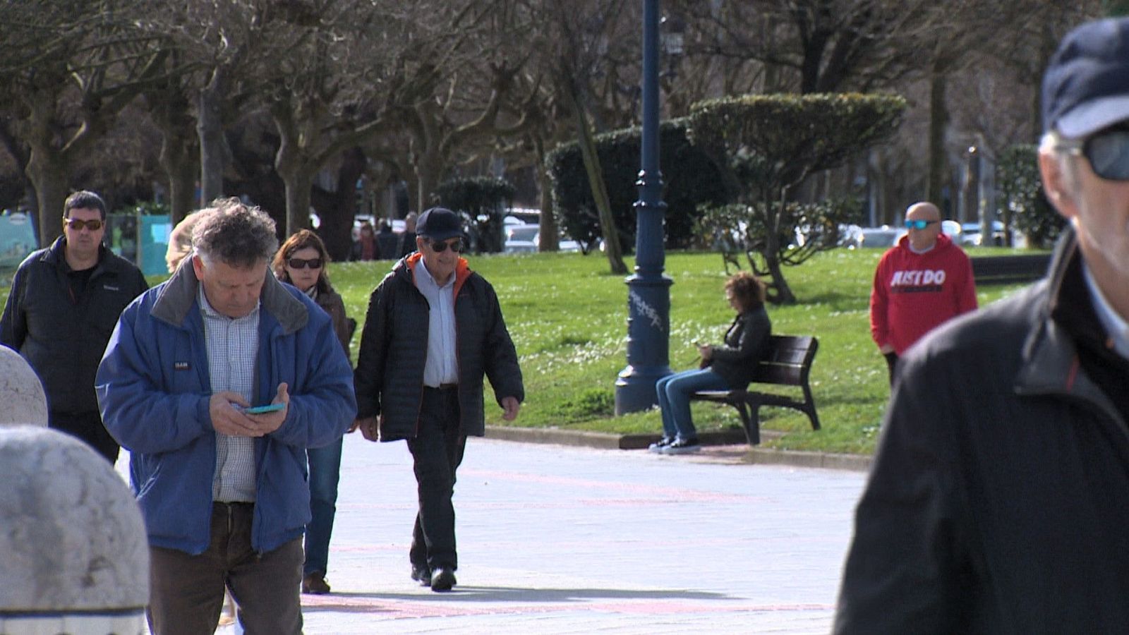 En la playa, un hombre con chaqueta azul y pantalones marrones mira su teléfono. Otros individuos caminan, incluyendo personas con gafas de sol y chaquetas oscuras. Al fondo, un hombre con sudadera roja y gafas de sol está de pie, y una mujer está sentada en un banco.