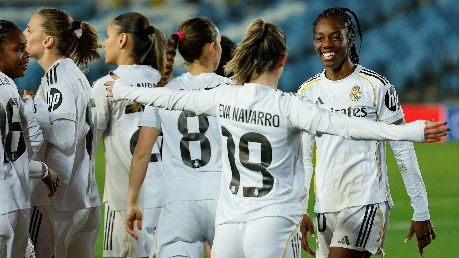 Jugadoras del Real Madrid celebran un gol en un campo de fútbol, vestidas con camisetas blancas. Se observa a Eva Navarro con el número 19 y a Naomie Feller sonriendo.