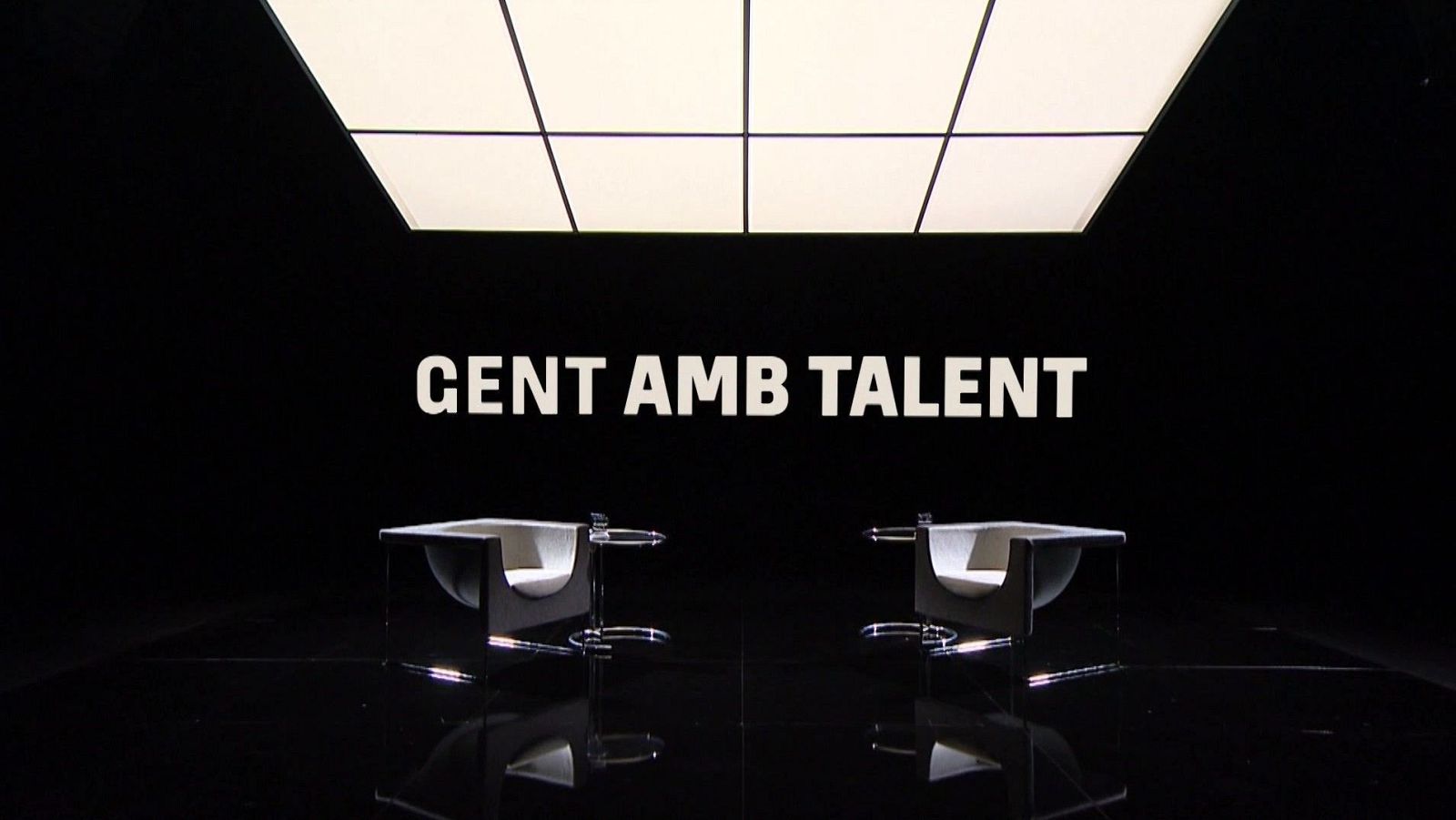 En un plató de televisió, dues cadires blanques i modernes es situen davant d'un fons negre amb el text "GENT AMB TALENT". El terra llustrós reflecteix la llum de l'escenari.