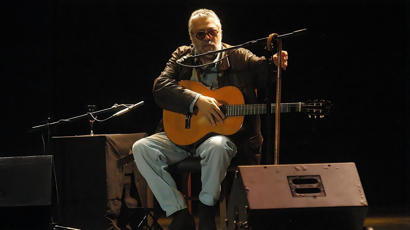 En un escenario con fondo negro, Facundo Cabral toca la guitarra, vestido con chaqueta de cuero y gafas redondas, frente a un micrófono y un atril con una copa de vino.