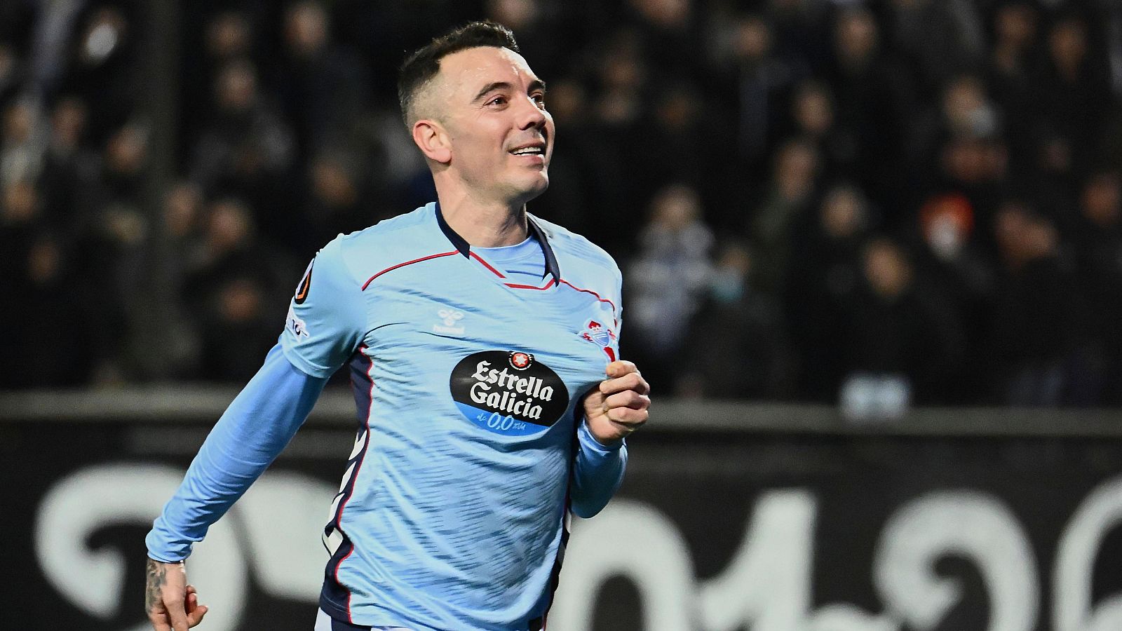 Un jugador de fútbol del Celta de Vigo, con la camiseta celeste y el logo de "Estrella Galicia", celebra un gol en un partido de la Europa League, con la afición en las gradas.