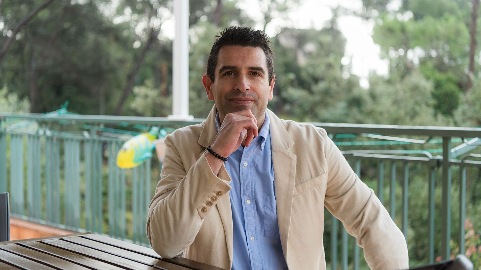 Sergio Marina, amb camisa blava clara i jaqueta crema, està assegut a una taula de fusta en un espai exterior, amb vegetació al fons.