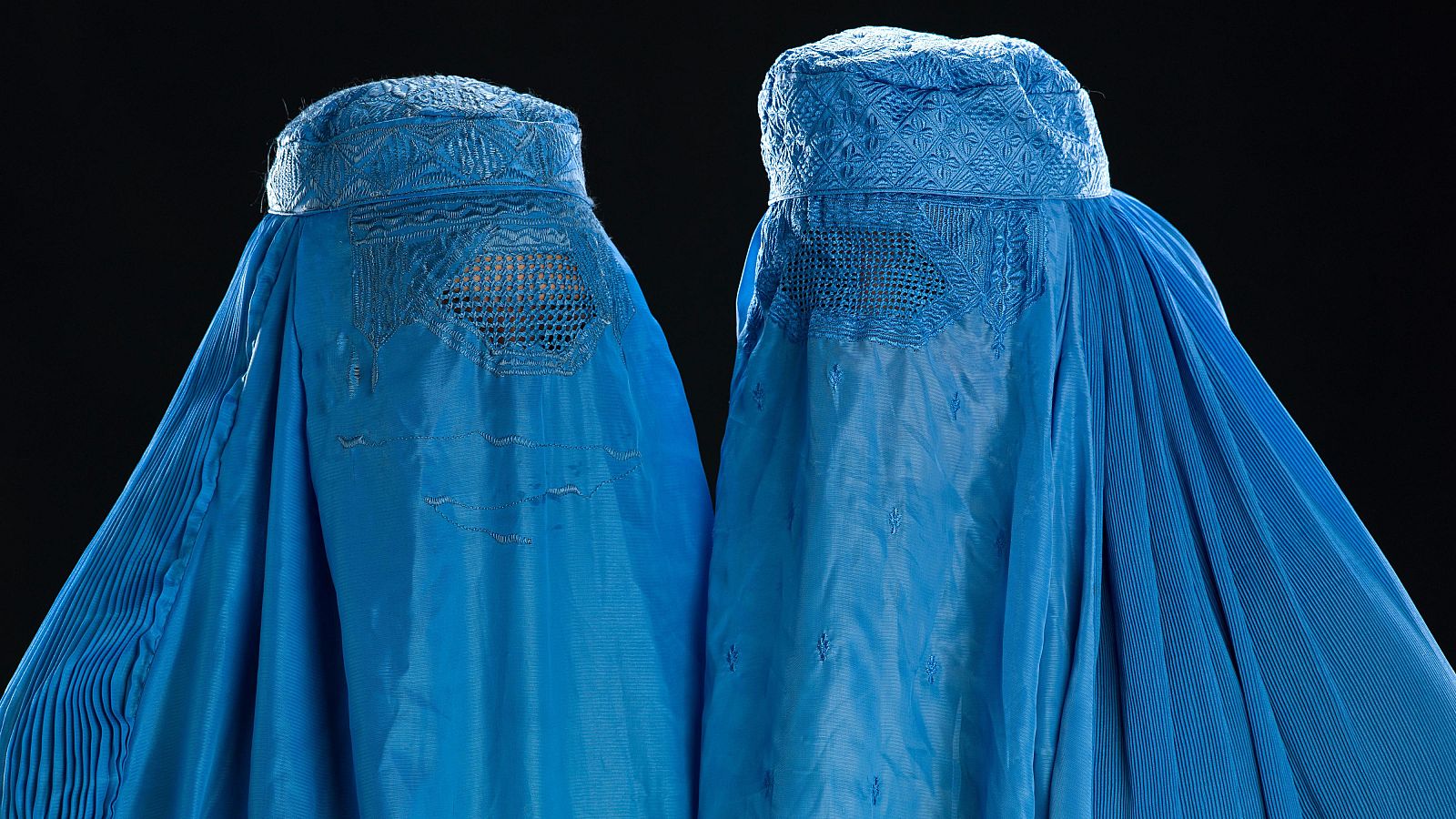 Dos burkas azules, con intrincados bordados en la parte superior, se alzan sobre un fondo negro, cubriendo por completo la figura femenina y dejando solo una rejilla para los ojos.