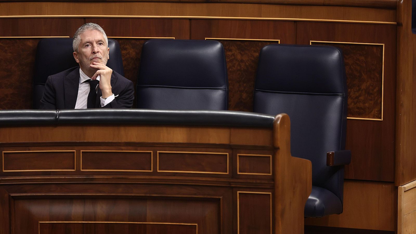 Fernando Grande-Marlaska, con traje oscuro y corbata, aparece pensativo sentado en un escaño de cuero en una sala con paneles de madera oscura y detalles dorados.
