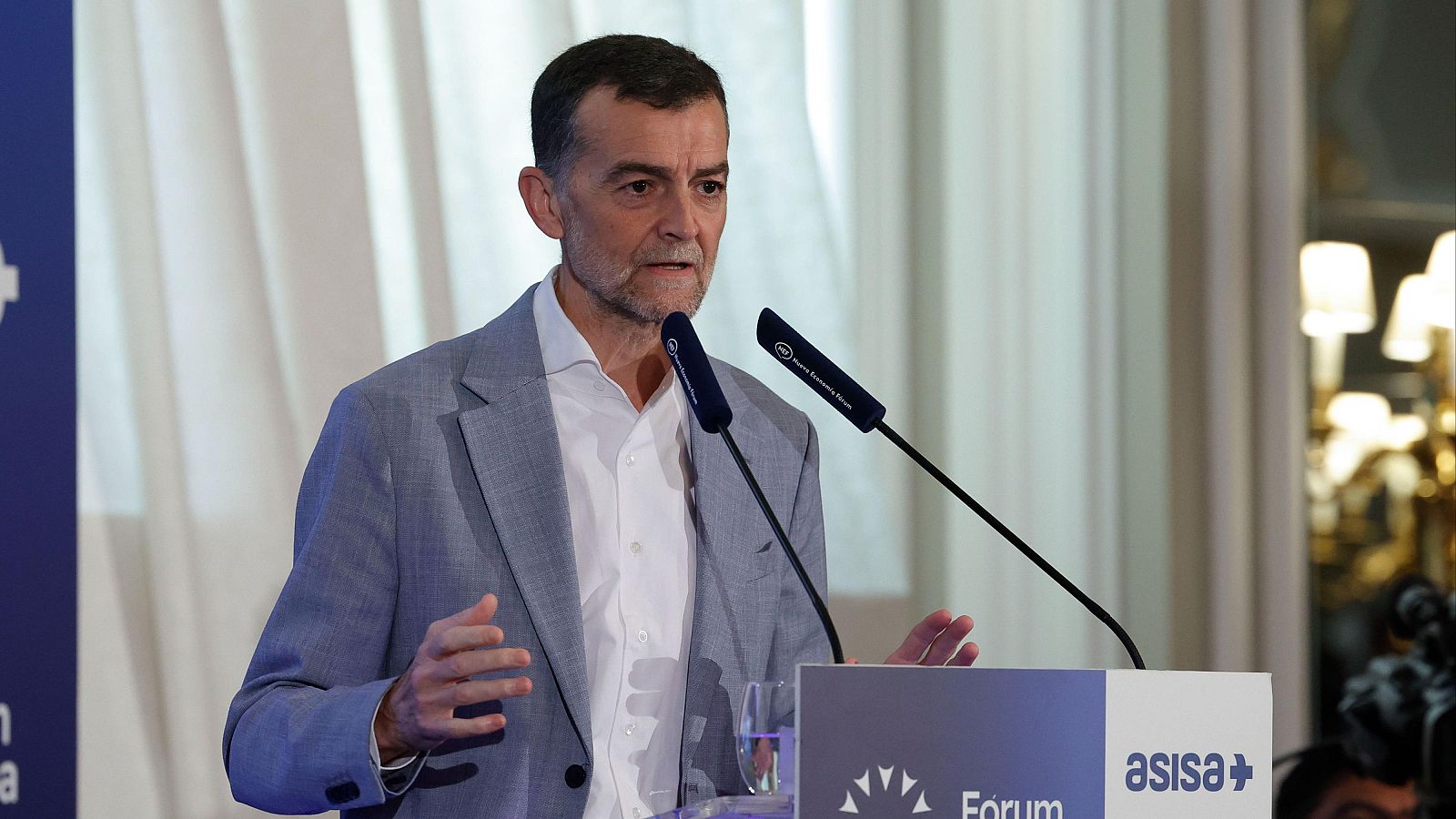 Antonio Maíllo, con camisa blanca y chaqueta clara, habla desde un atril con logos, posiblemente en un evento informativo. Se observan micrófonos y una cortina blanca al fondo.