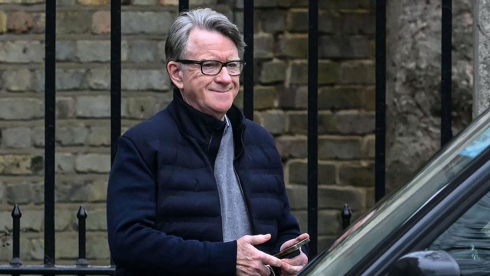 Peter Mandelson, con chaqueta azul marino y gafas, sonríe mientras sostiene un teléfono móvil en una calle urbana, con una pared de ladrillo y una verja como fondo. La imagen fue tomada en febrero de 2026.