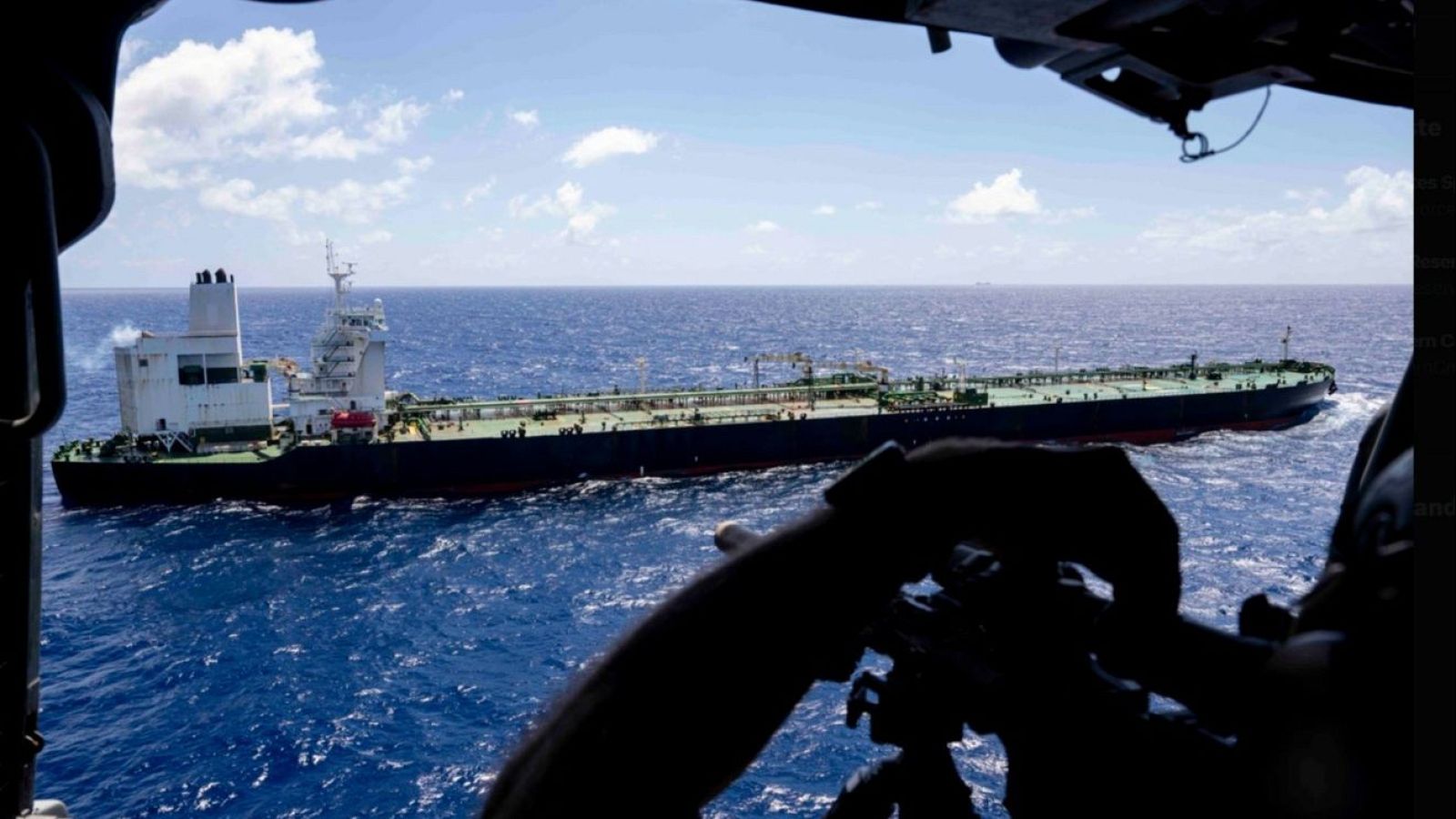 Un gran petrolero oscuro navega en un mar azul, con una estructura blanca visible. El cielo, con nubes blancas, sugiere un día soleado. La imagen fue tomada desde un helicóptero, cuya silueta se aprecia en primer plano.