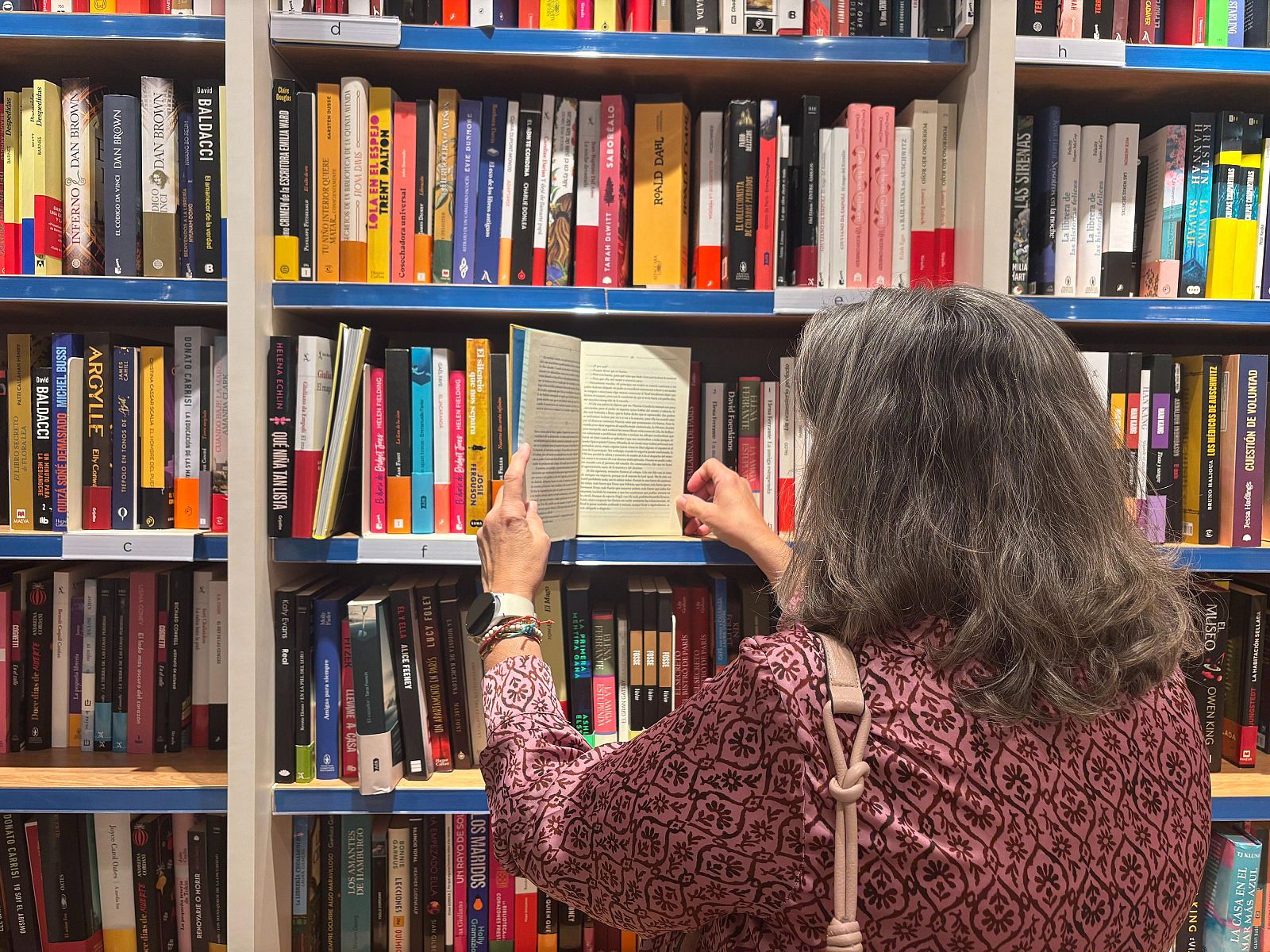 Una mujer observa un libro abierto frente a una estantería repleta de ejemplares de diversos tamaños y colores. Se distingue una blusa estampada y una bandolera clara. Entre los títulos visibles, se encuentran obras de Dan Brown y Baldacci.