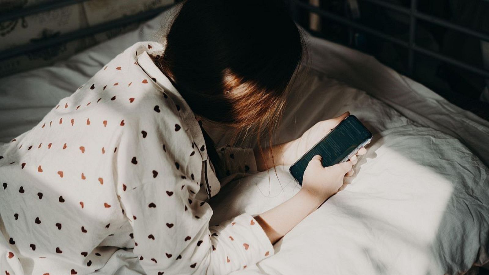 Un menor, vestido con pijama de corazones, yace en una cama blanca, absorto en la pantalla de su teléfono móvil en una habitación oscura.