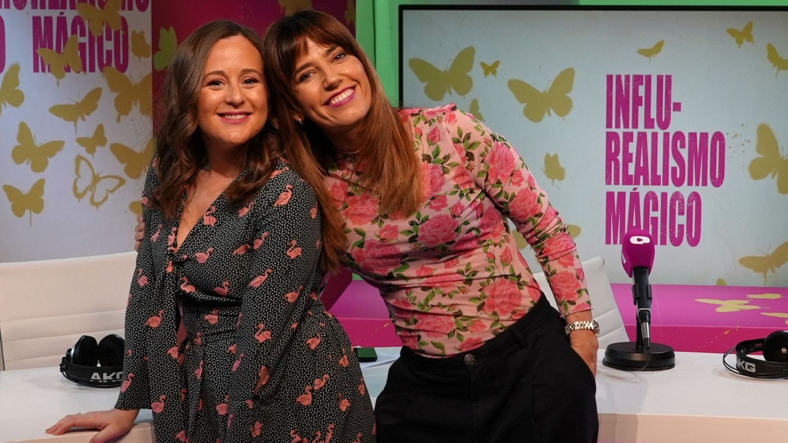 Dos mujeres sonríen en un estudio de radio. Una de ellas viste un vestido negro con estampado, la otra una blusa estampada y pantalones oscuros. Detrás, un fondo con letras rosas y mariposas doradas.