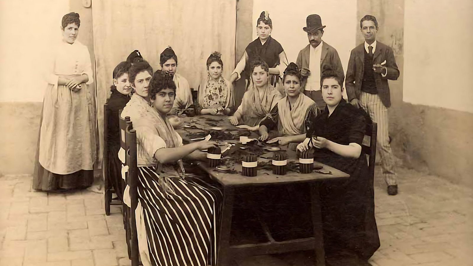 En una fábrica de tabacos, un grupo de mujeres, vestidas con ropa de época, trabajan en la elaboración de cigarros alrededor de una mesa. Dos hombres observan la escena, mientras que hojas de tabaco y cigarros ya hechos se encuentran sobre la mesa.