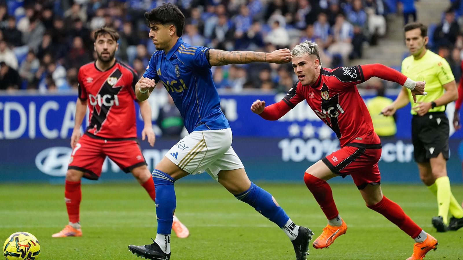 Un jugador del Oviedo, con camiseta azul, avanza con el balón en un partido de fútbol, mientras dos rivales vestidos de rojo intentan detenerlo. El árbitro, de amarillo y negro, está atento a la jugada, con el público de fondo.