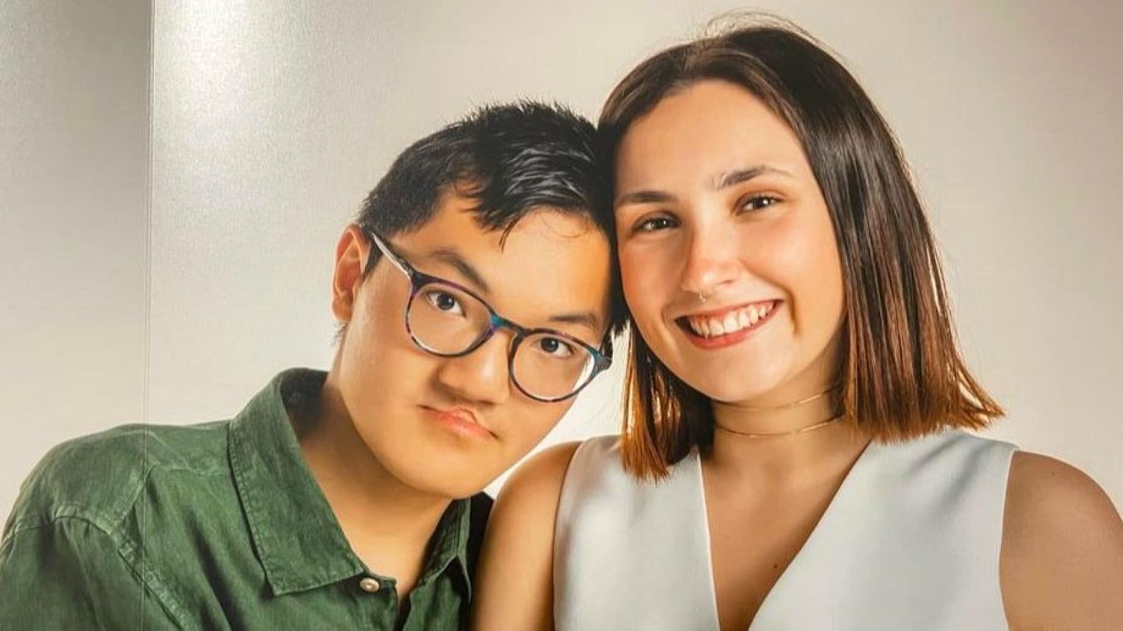 Dos jóvenes sonríen y posan juntos; uno con gafas y camisa verde, el otro con un top blanco, muy cerca el uno del otro, con un fondo claro.