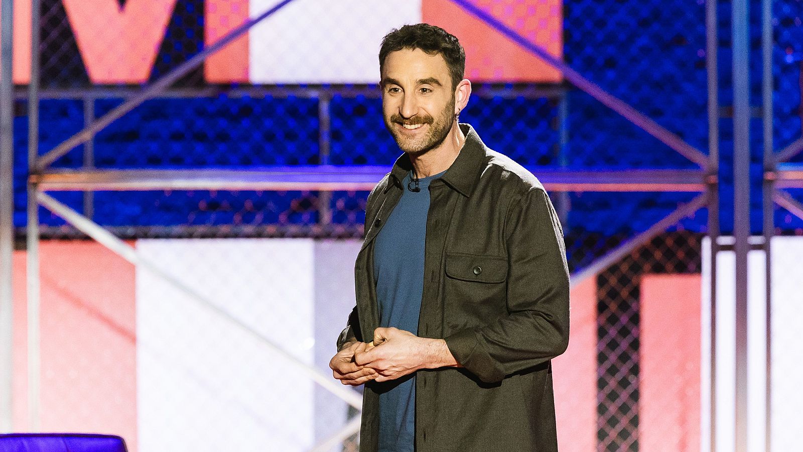 En la imagen, Dani Rovira sonríe con las manos entrelazadas, luciendo una camiseta azul y una chaqueta verde oscuro, frente a un fondo decorativo con formas geométricas.