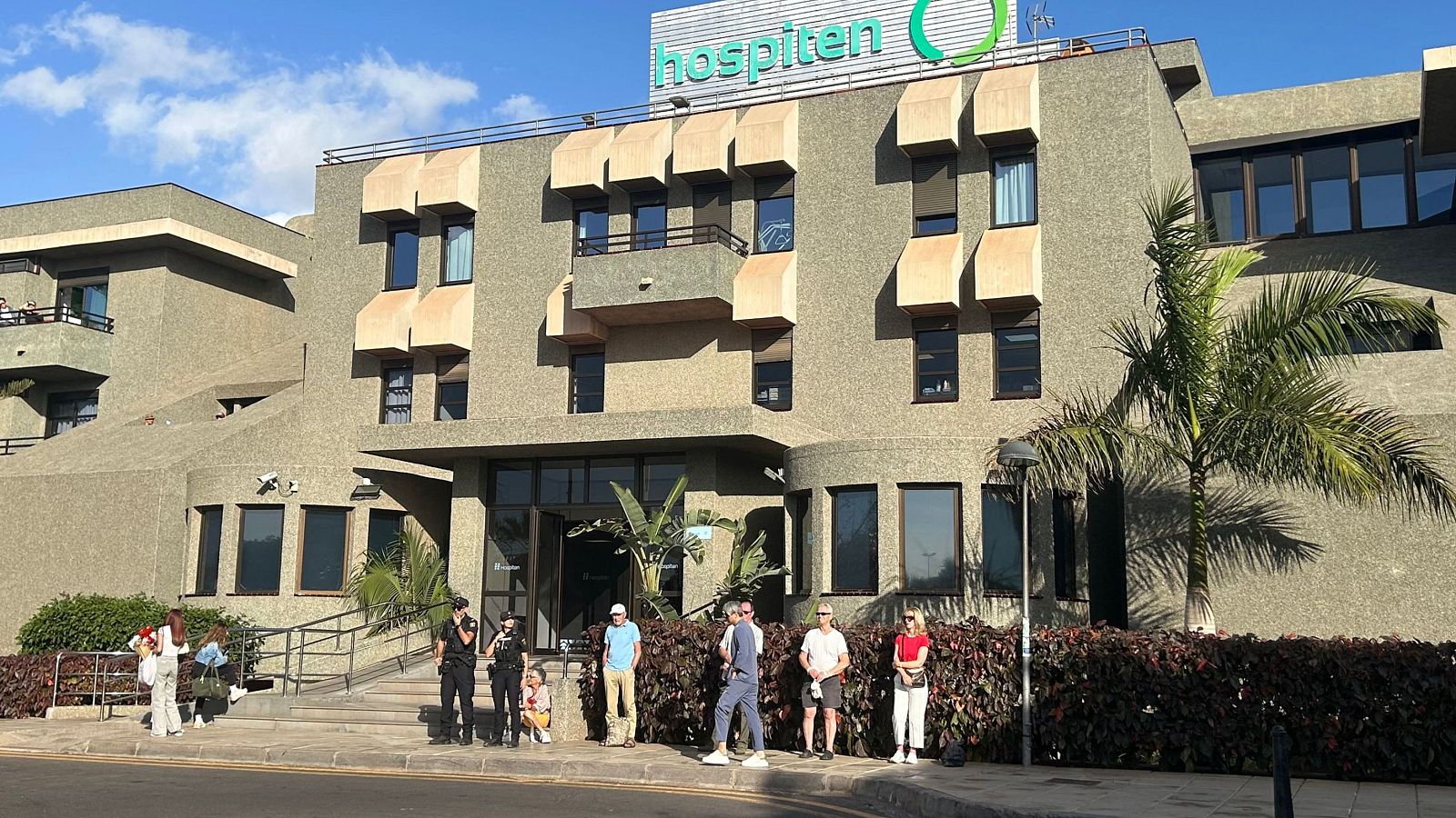 Un edificio moderno de color grisáceo, con el letrero "Hospiten" en azul, se alza con múltiples ventanas. Frente al edificio, un grupo de personas vestidas de forma casual se encuentran en la acera.