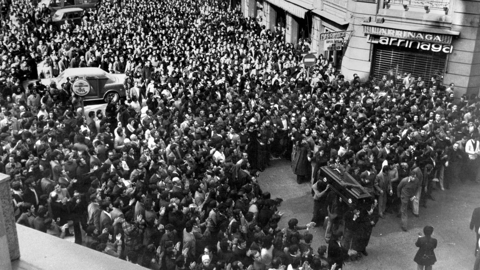 Una imagen en blanco y negro muestra una calle llena de gente en un ambiente solemne. En el centro, un grupo de hombres transportan féretros, mientras que la multitud los rodea. Se observan edificios con letreros.