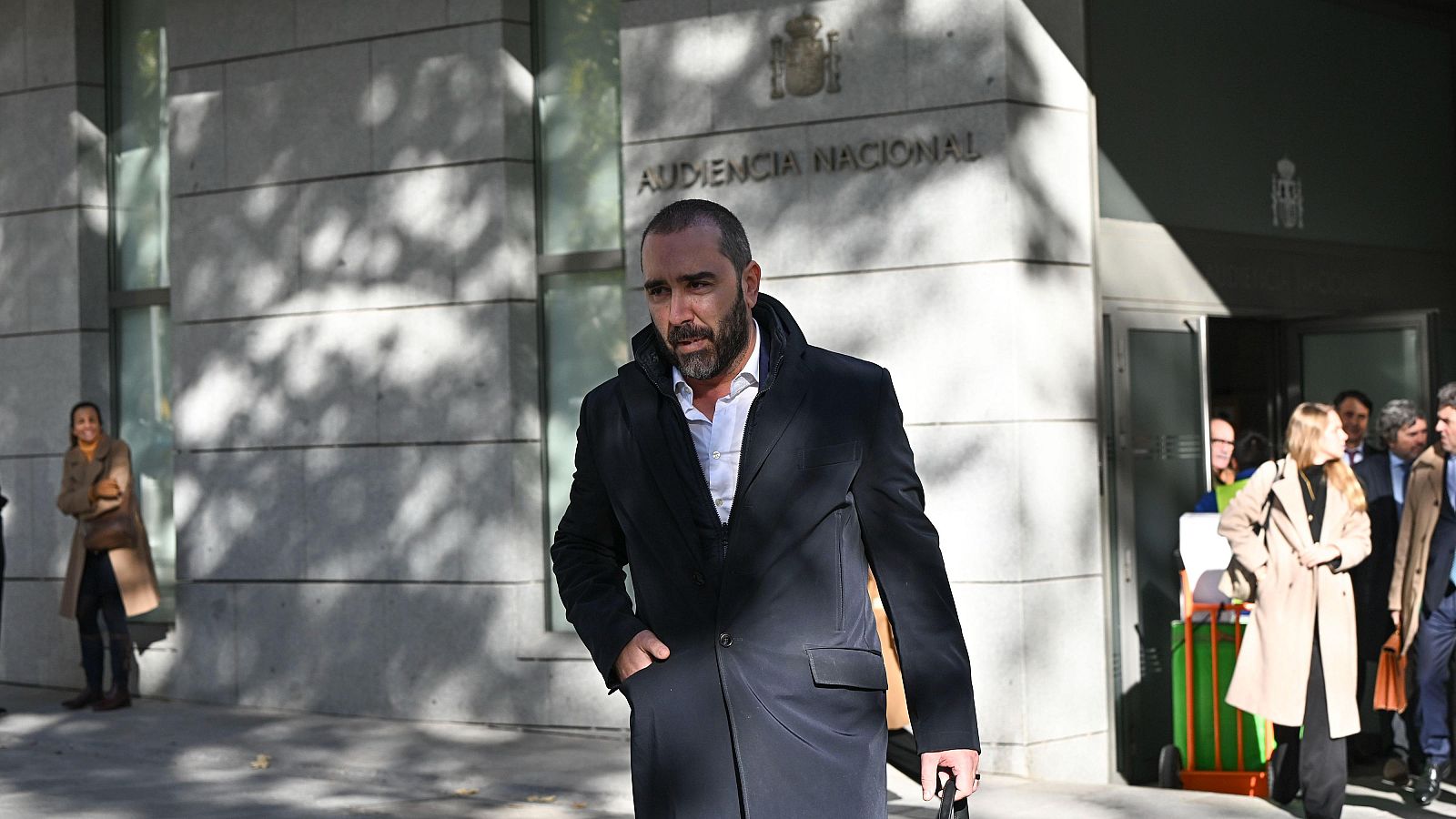 Un hombre con barba y abrigo oscuro, posiblemente De Aldama, sale de un edificio con el nombre "Audiencia Nacional" visible, con una expresión seria y una mano en el bolsillo.