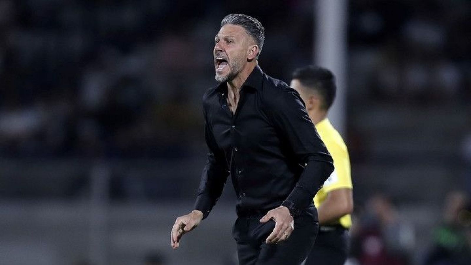 Martín Demichelis, con camisa negra y pantalones oscuros, gesticula y grita en un campo de fútbol, posiblemente durante un partido. Se observan otras personas y gradas al fondo.