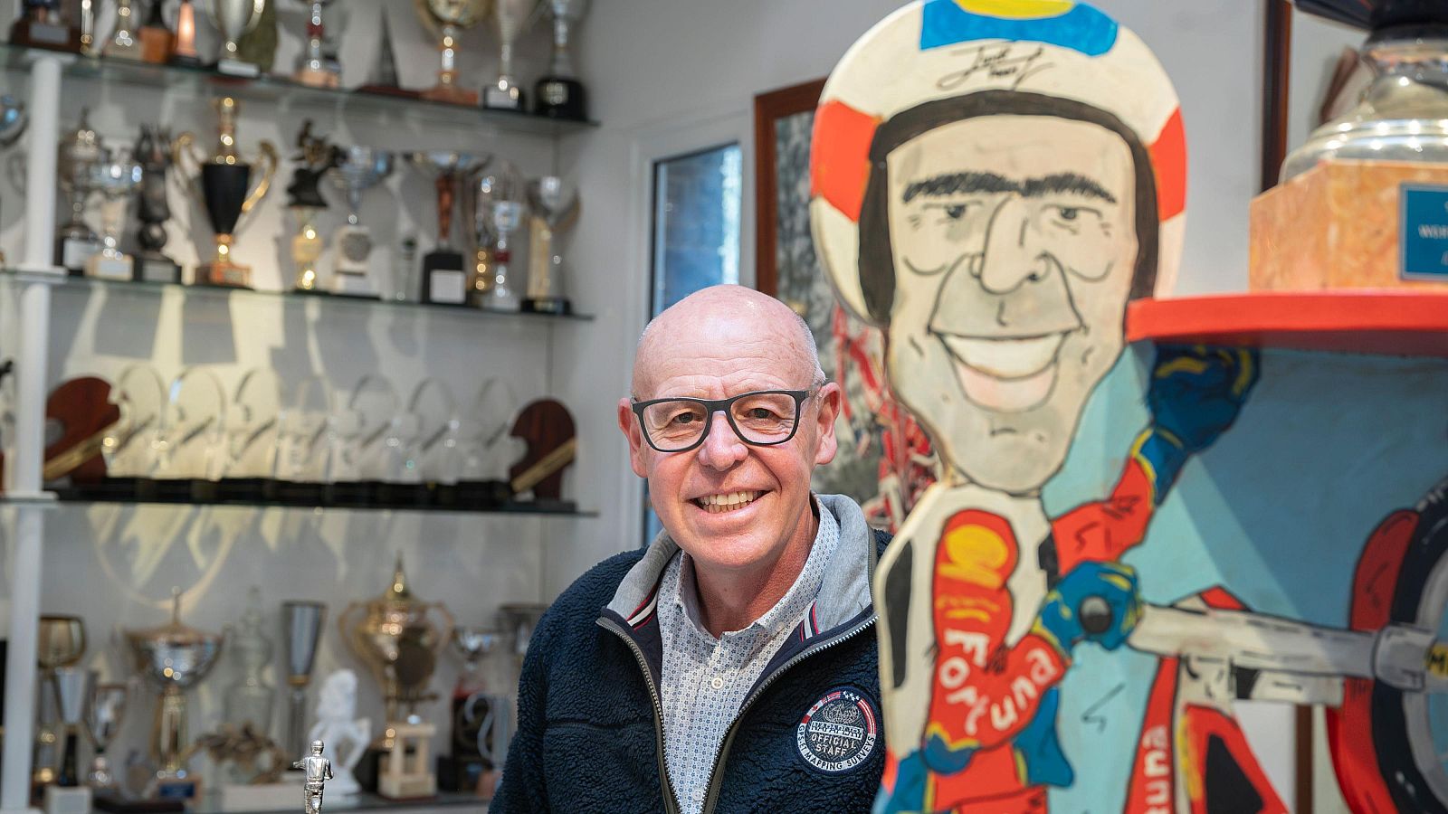 Jordi Tarrés, amb ulleres i jaqueta blava, somriu davant la càmera. Darrere seu, una vitrina plena de trofeus i una caricatura d'un pilot de trial sobre una moto.