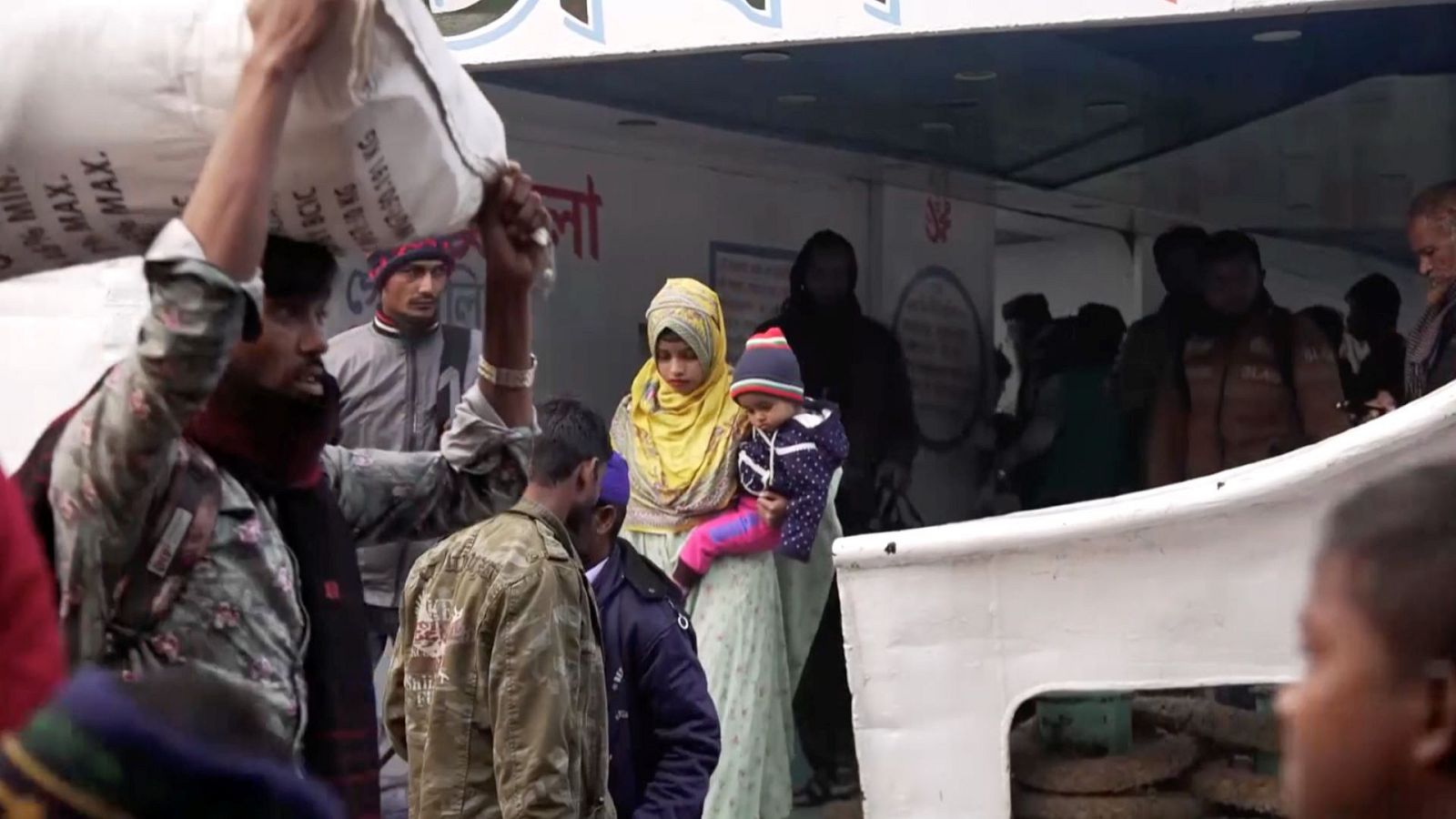 Un grupo de personas en Bangladés, algunos cargando sacos grandes, posiblemente de ayuda, y una mujer con un niño en brazos, se preparan para un traslado, posiblemente en un vehículo.