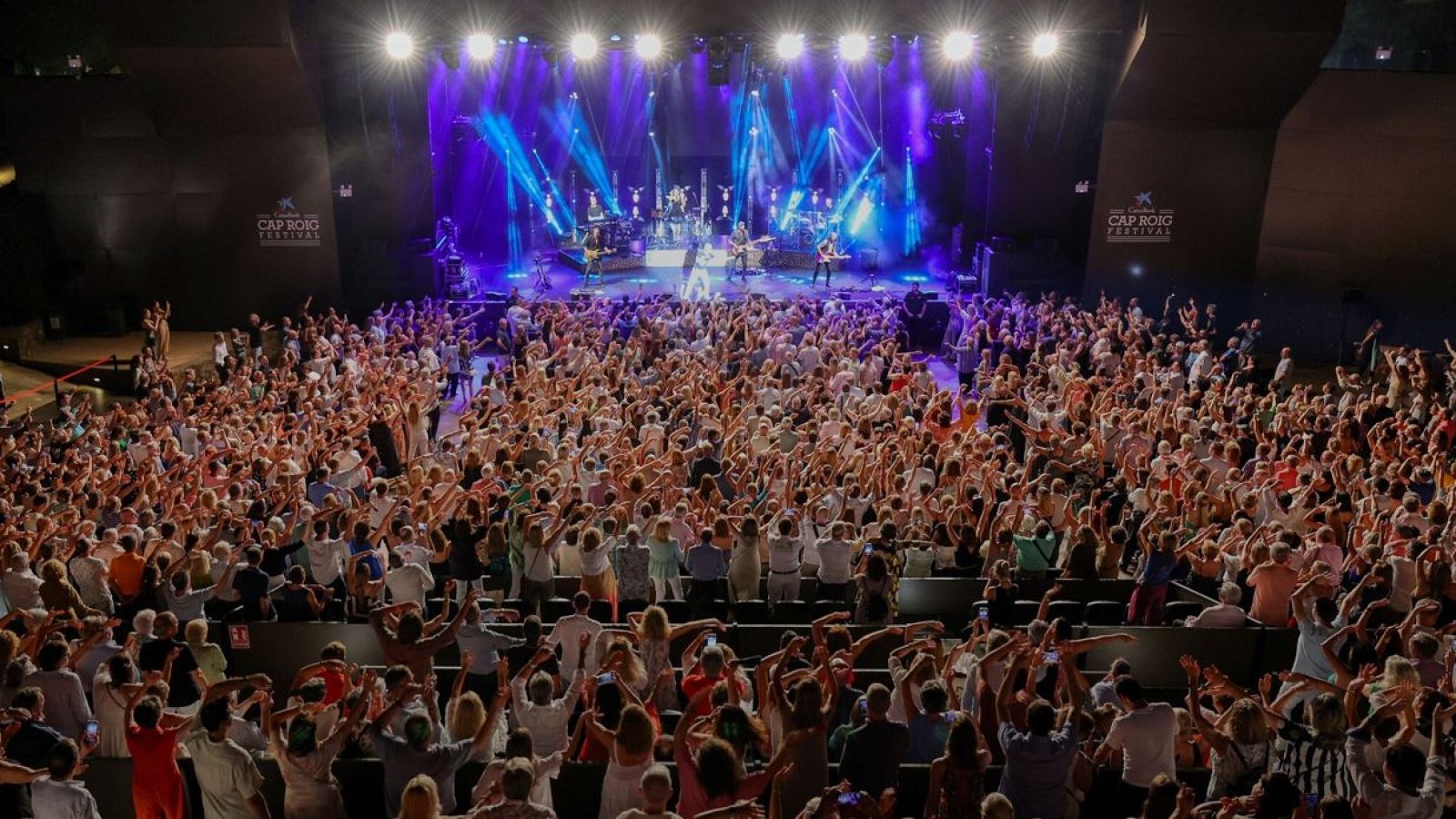 En el escenario, una banda musical se presenta ante una multitud entusiasta, bajo la intensa iluminación de focos. El ambiente festivo sugiere un evento musical vibrante y lleno de energía.