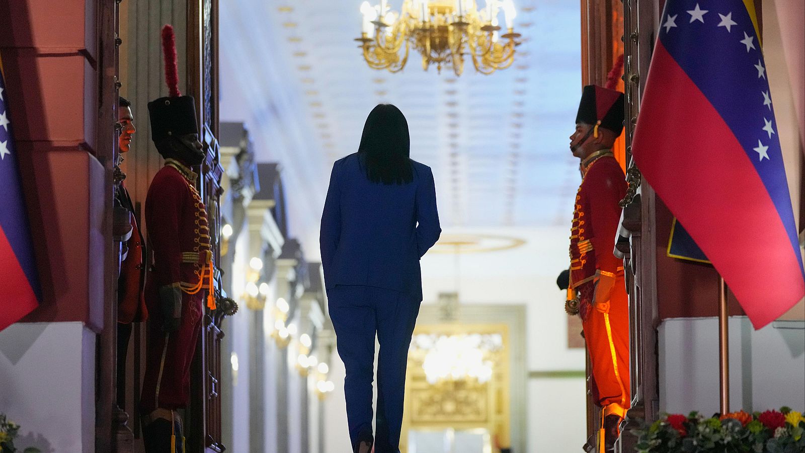 En el Palacio de Miraflores, una mujer con traje oscuro se dirige hacia el interior, custodiada por guardias uniformados. El entorno se caracteriza por detalles dorados y una bandera.